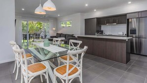 Uma cozinha moderna com armários escuros e eletrodomésticos de aço inoxidável, uma mesa de jantar de vidro com cadeiras brancas e uma grande janela com vista para o verde.