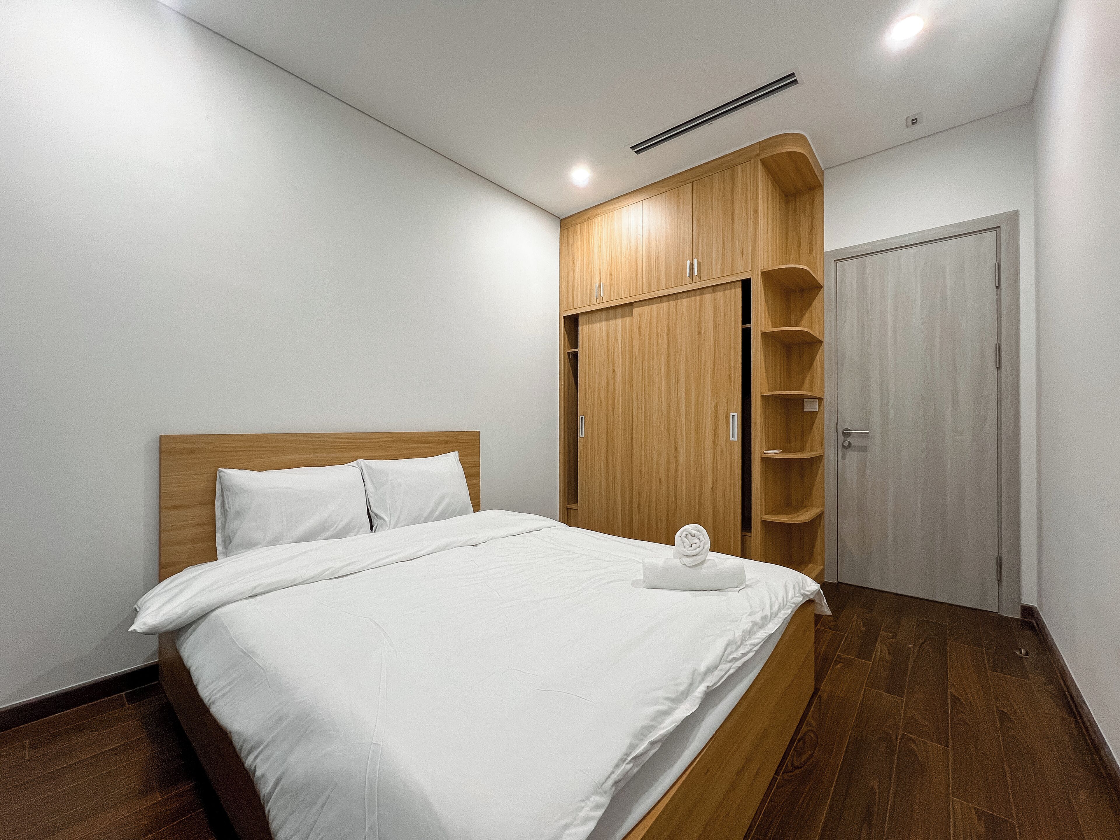 A bedroom with a wooden bed frame, white bedding, a wardrobe with shelves, and a door.