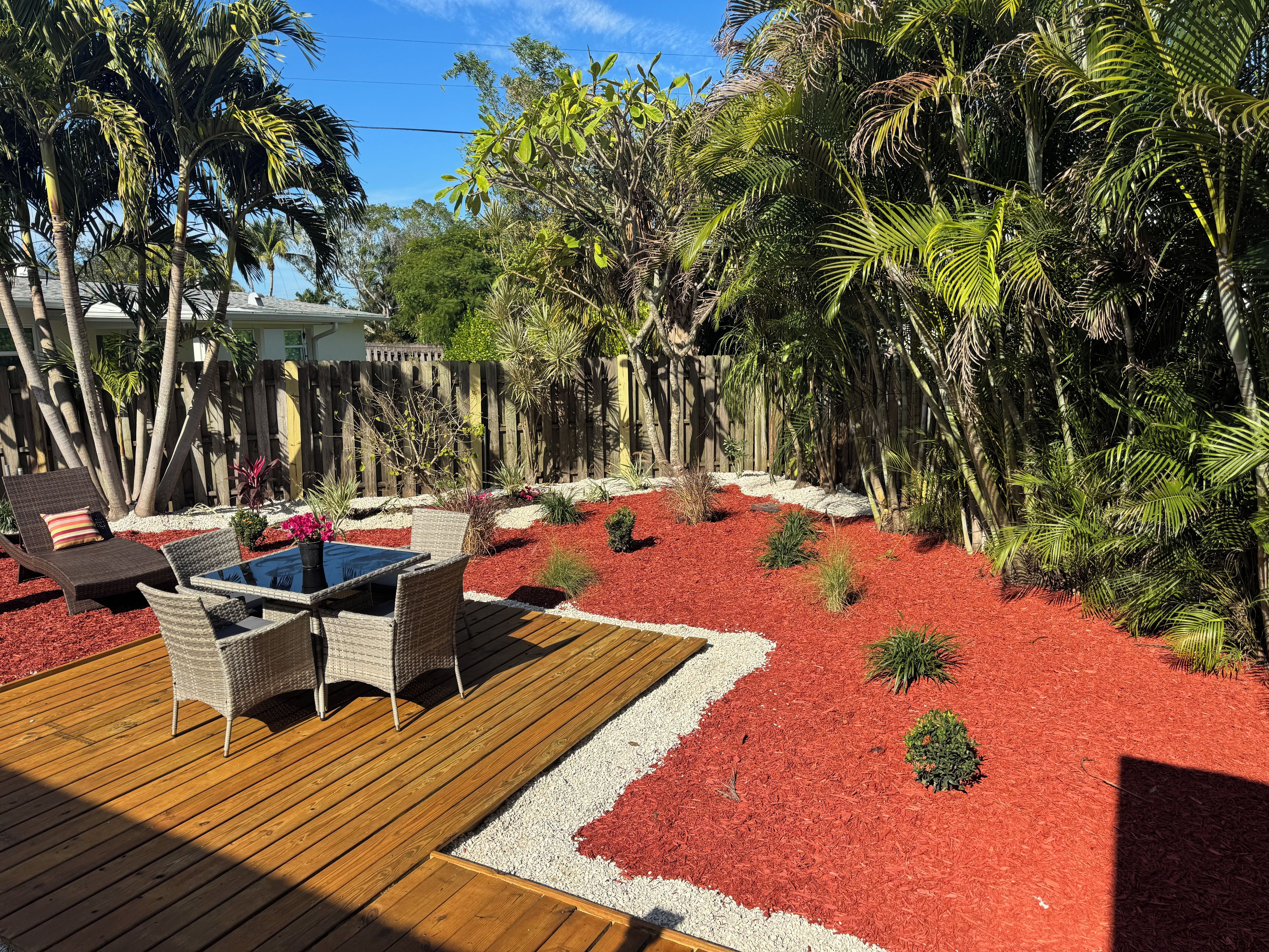 Une terrasse en bois avec des meubles d’extérieur et un espace jardin agrémenté de paillis rouge et de différentes plantes.