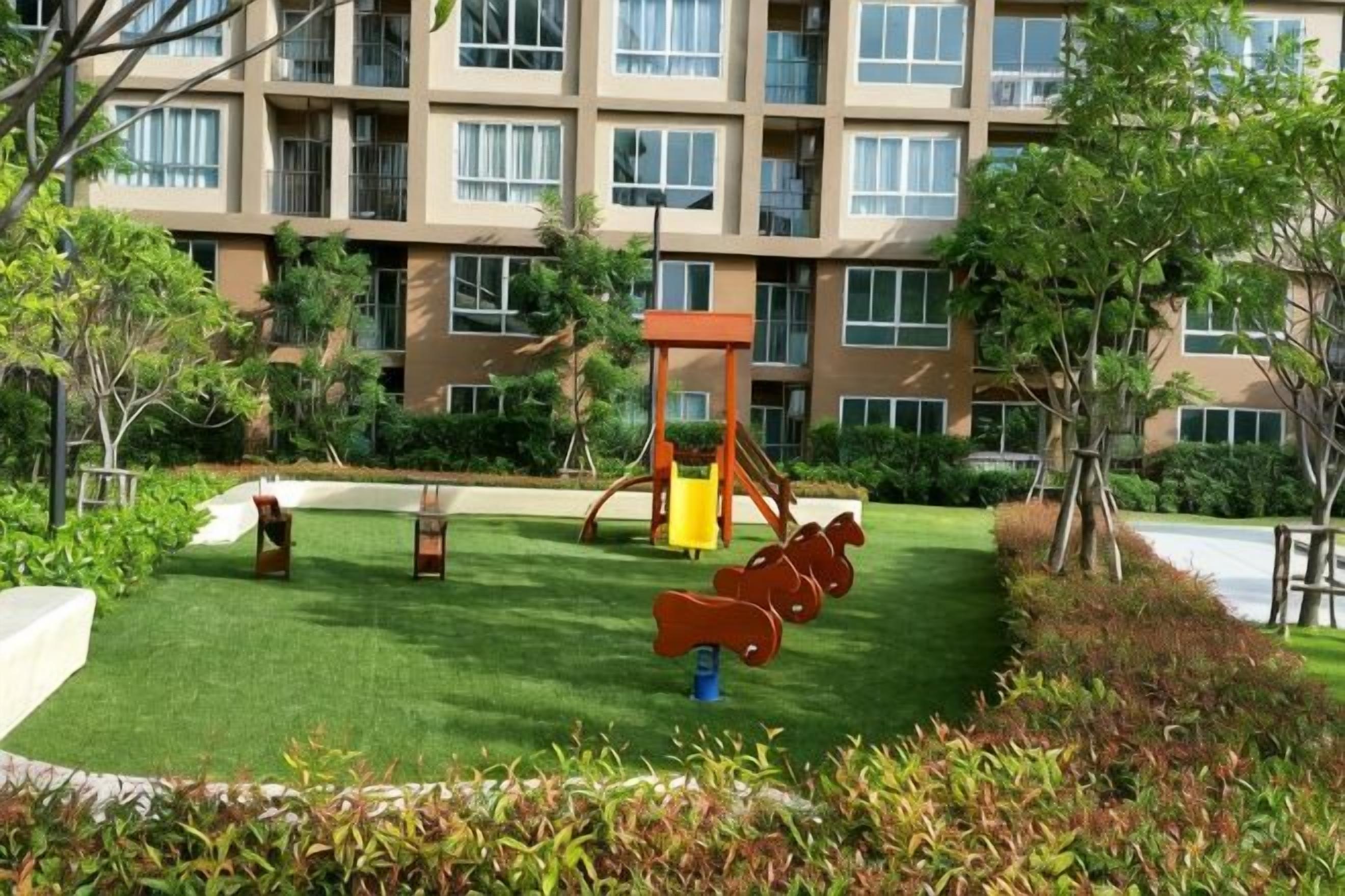 Un parque infantil con columpios y tobogán, rodeado de un césped cuidado y árboles, frente a un edificio de varios pisos.