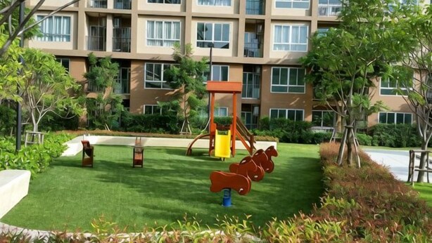 A playground with swings and a slide, surrounded by a well-maintained lawn and trees, in front of a multi-story building. - The Relaxing Room Mountain View at Baan Tew Lom Condominium Cha am - Hua Hin (Cha-am)