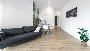 A modern living room with a dark grey sofa, a dining table with chairs, a potted plant, and a cityscape painting on the wall.