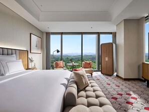 A hotel room with a large bed, a sofa, a small table, and a view of the mountains. - Swissotel Nusantara (Bukit Subur)