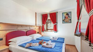 Une chambre d’hôtel avec une tête de lit rouge, deux lits avec une literie bleue, une table de chevet en bois, un radiateur et une fenêtre avec des rideaux rouges.