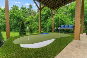 A covered putting green with a swing chair, surrounded by lush greenery.