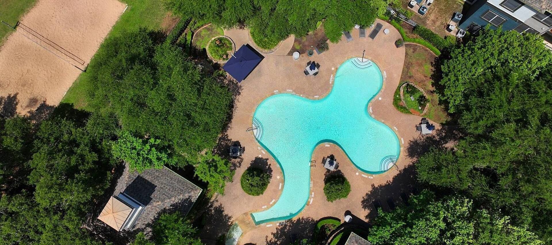 An aerial view of a swimming pool with a unique shape, surrounded by greenery and a few seating areas.