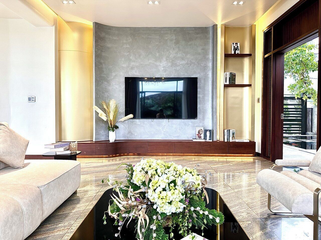 A modern living room with a large flat-screen TV mounted on a textured wall, two sofas, a coffee table with flowers, and a bookshelf with books and decorative items.