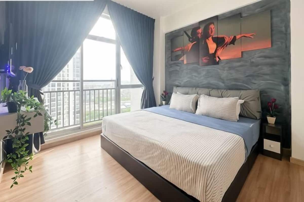 A bedroom with a large bed, a desk with a plant, a window with curtains, and a city view.