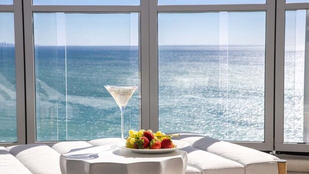 Ein modernes Zimmer mit großem Fenster zum Meerblick, einer weißen gepolsterten Sitzecke und einem Tisch mit Früchten und einem Martini-Glas.