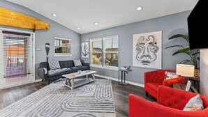 Un salon moderne avec un canapé gris, un fauteuil rouge et une table basse. De grandes fenêtres sont équipées de stores, une planche de surf est accrochée au mur, et des œuvres d’art ornent les murs.