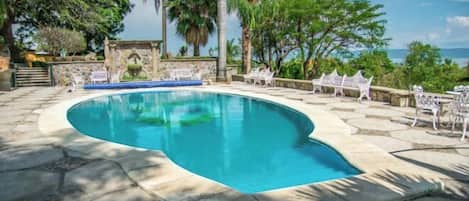 A swimming pool with a curved edge, surrounded by stone tiles and white chairs.