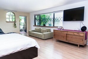 Un dormitorio con cama, sofá, cómoda de madera, televisor y ventana con vistas a la vegetación.