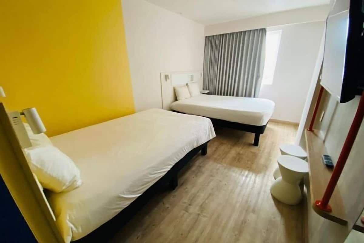 A hotel room with two beds, a small round table, a toilet, and a window with curtains.