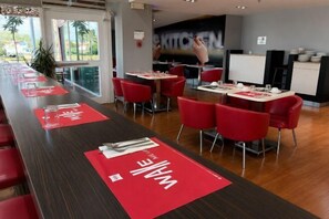 A modern dining area with red chairs and tables, a long counter, and a view of the outdoors. - Ibis Budget Aguascalientes (Aguascalientes)