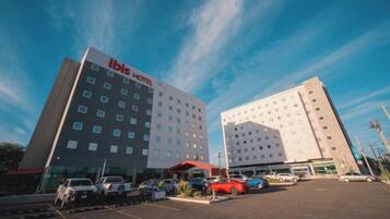 A hotel with a modern design, featuring a prominent 'ibis HOTEL' sign, a parking lot with several cars, and a clear blue sky in the background.