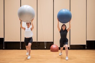 Zwei Personen führen Übungen mit großen Gymnastikbällen in einem Raum mit Holzboden und beigen Wänden aus.