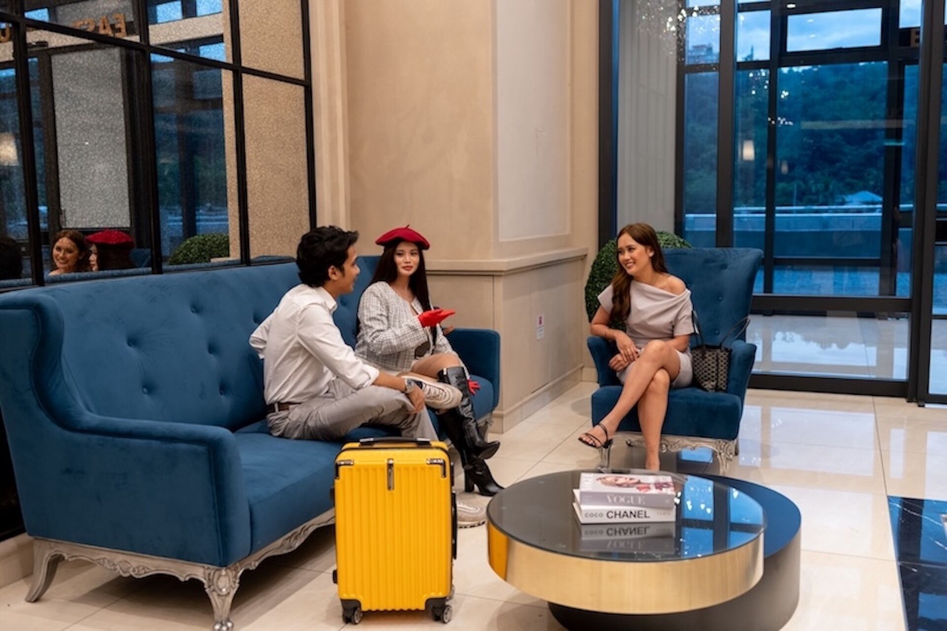 A modern lobby with a blue sofa, a yellow suitcase, and a round glass coffee table with a book.