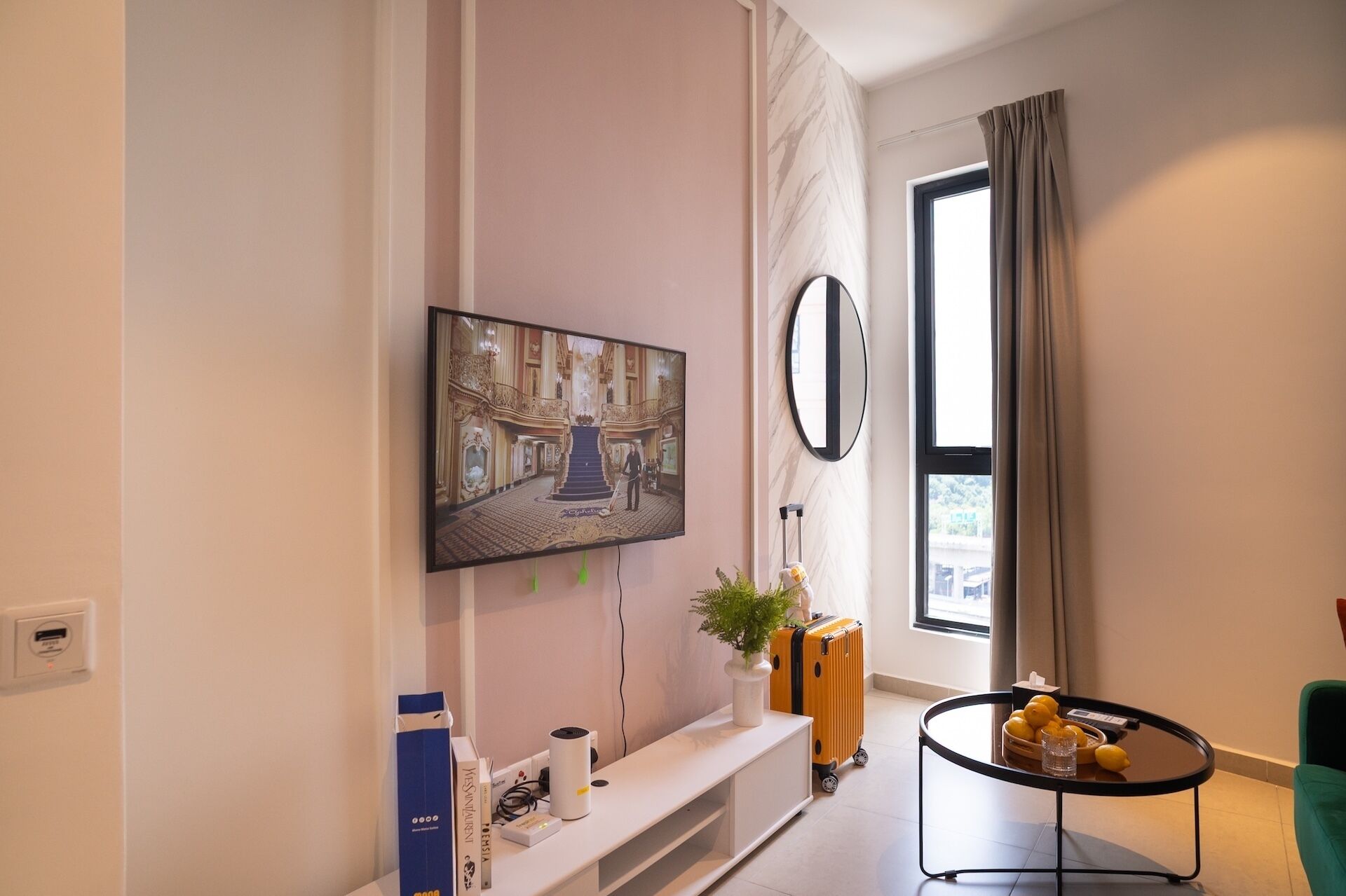 A modern living room with a flat-screen TV mounted on the wall, a round mirror, a small round table with a glass, and a bench.