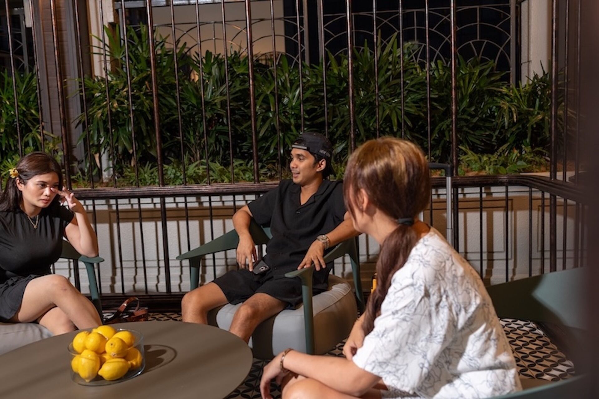 Three people seated on outdoor furniture with a table in front of them, surrounded by greenery and a metal fence.