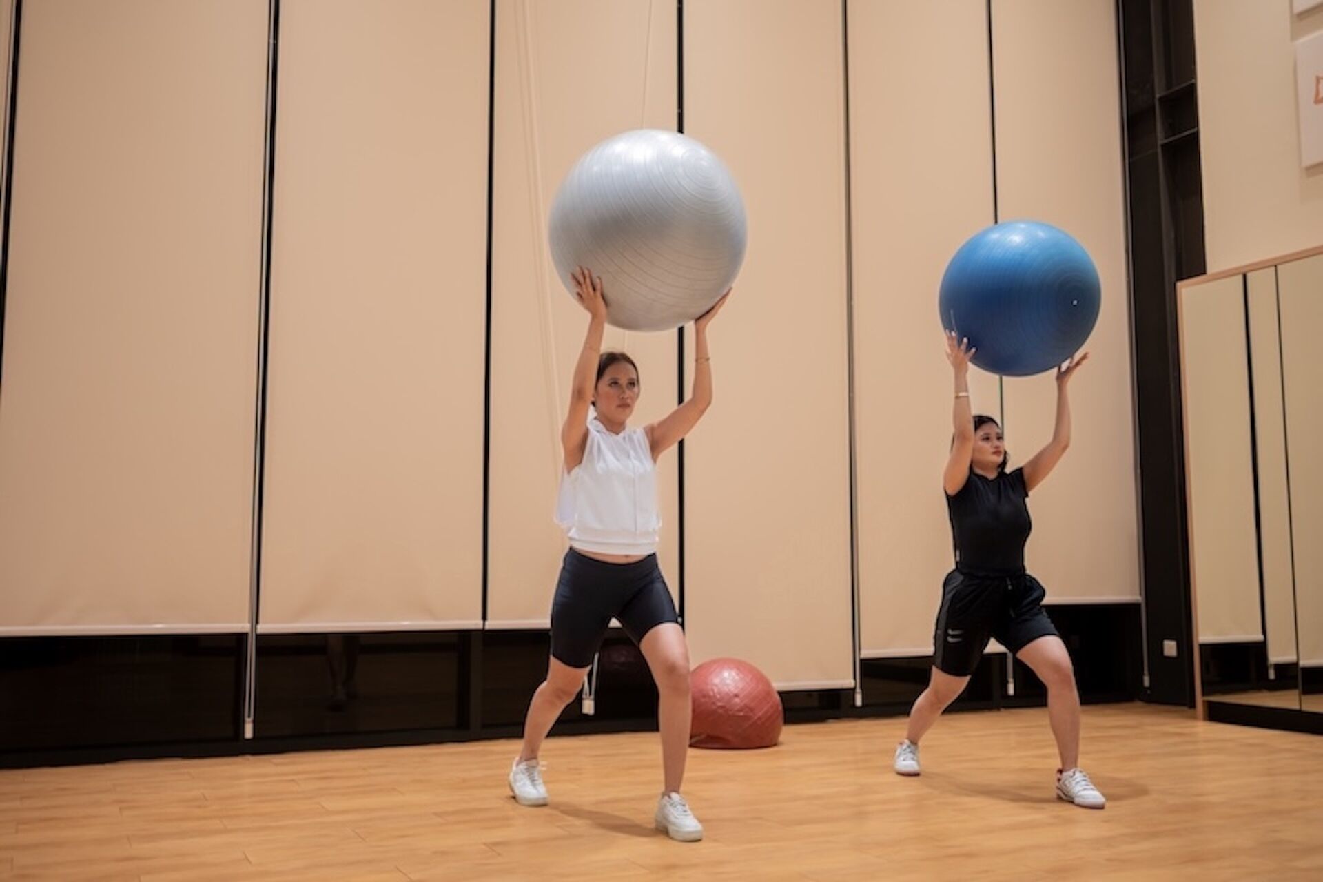 Two individuals exercising with large exercise balls in a room with a wooden floor and a large mirror on one wall.