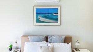 Ett välordnat sovrum med en säng, kuddar, en inramad bild av en strand, sängbord med lampor och en väggmonterad luftkonditioneringsanläggning.