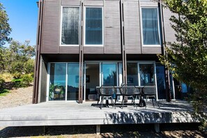 Une maison moderne à deux étages, dotée de grandes fenêtres en verre et d’une terrasse avec coin salon extérieur.
