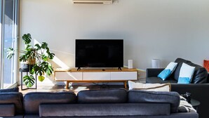 Ett modernt vardagsrum med en platt-TV placerad på en träkonsol, en svart lädersoffa och en stor krukväxt.
