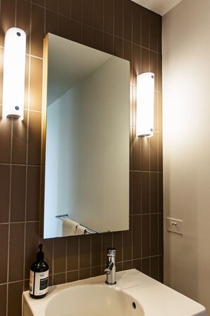 Une salle de bain avec un grand miroir, deux appliques murales, un lavabo et un flacon de savon liquide.