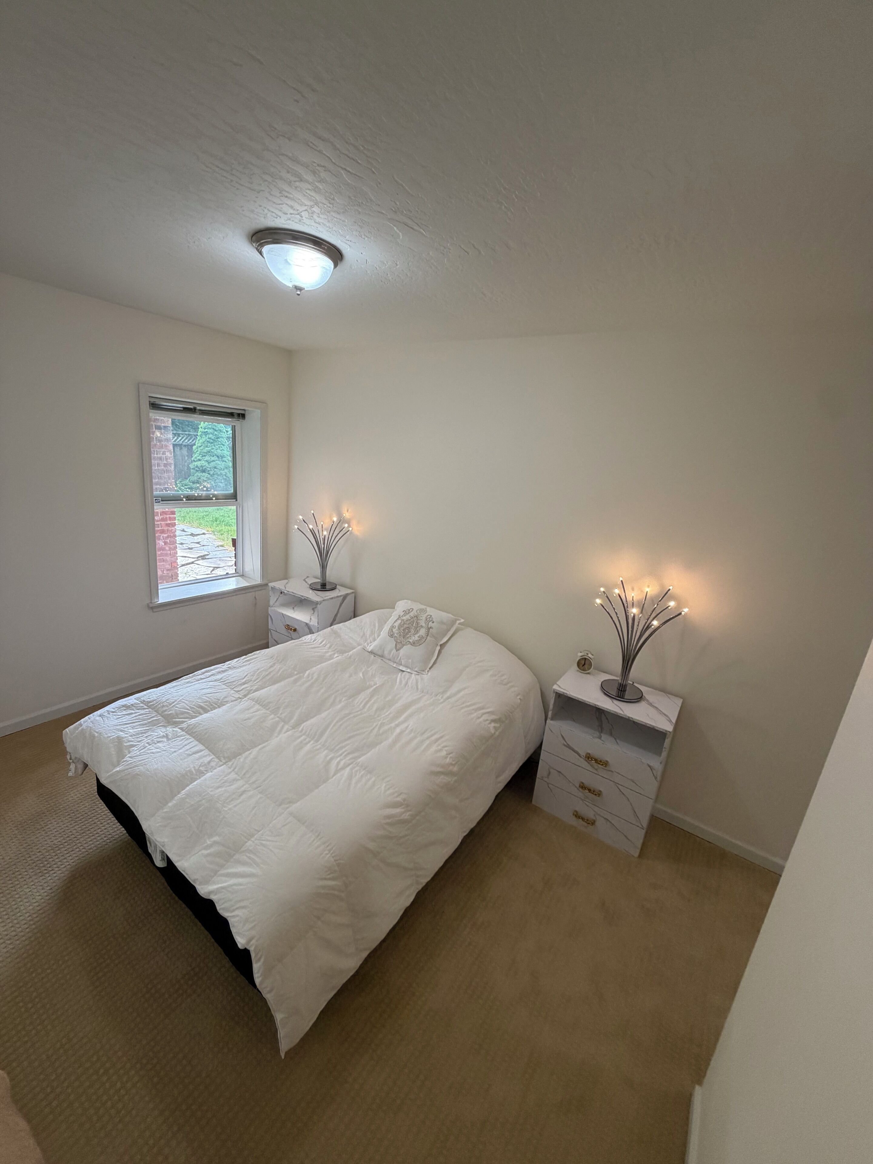 A bedroom with a bed, a nightstand, a dresser, a window, and a ceiling light.