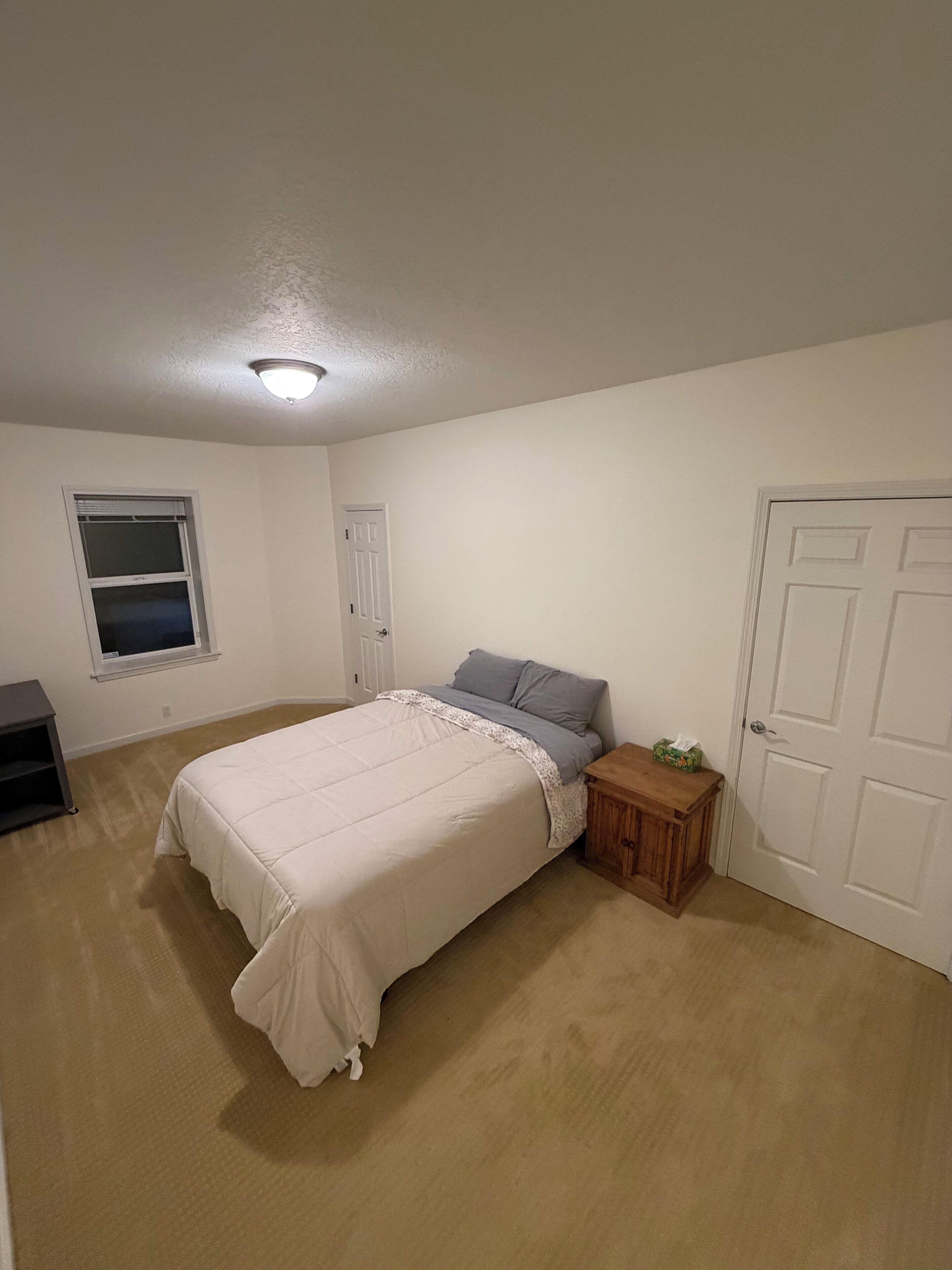 A bedroom with a bed, a nightstand, a window, a door, and a small shelf.