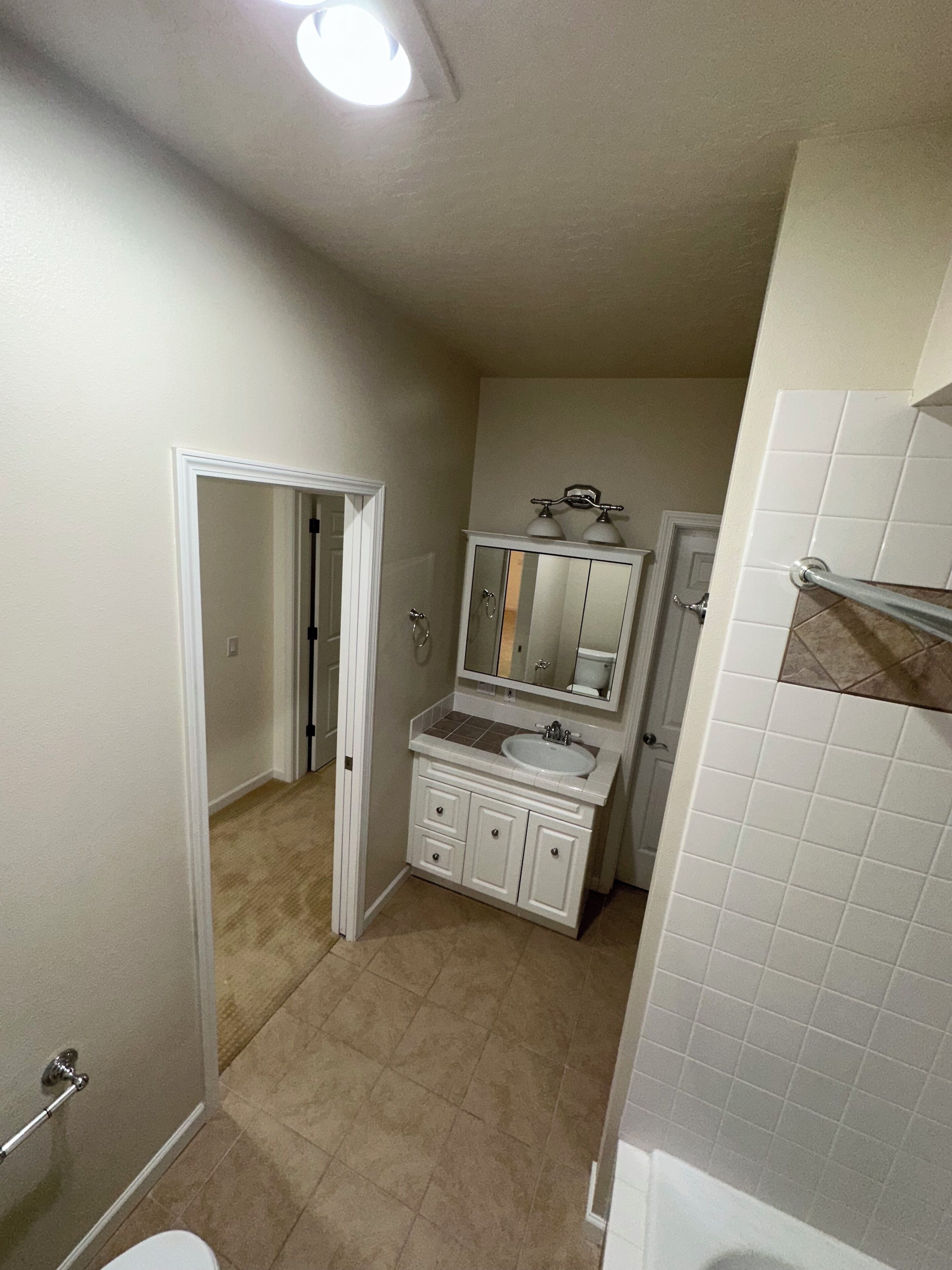 A bathroom with a white sink, mirror, and vanity, beige tiles, and a door leading to another room.