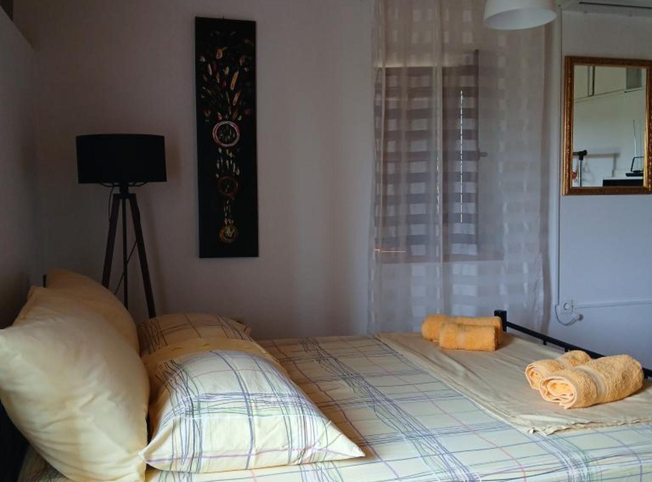 Quarto com cama, almofadas, luminária, pintura, janela com cortinas e espelho.