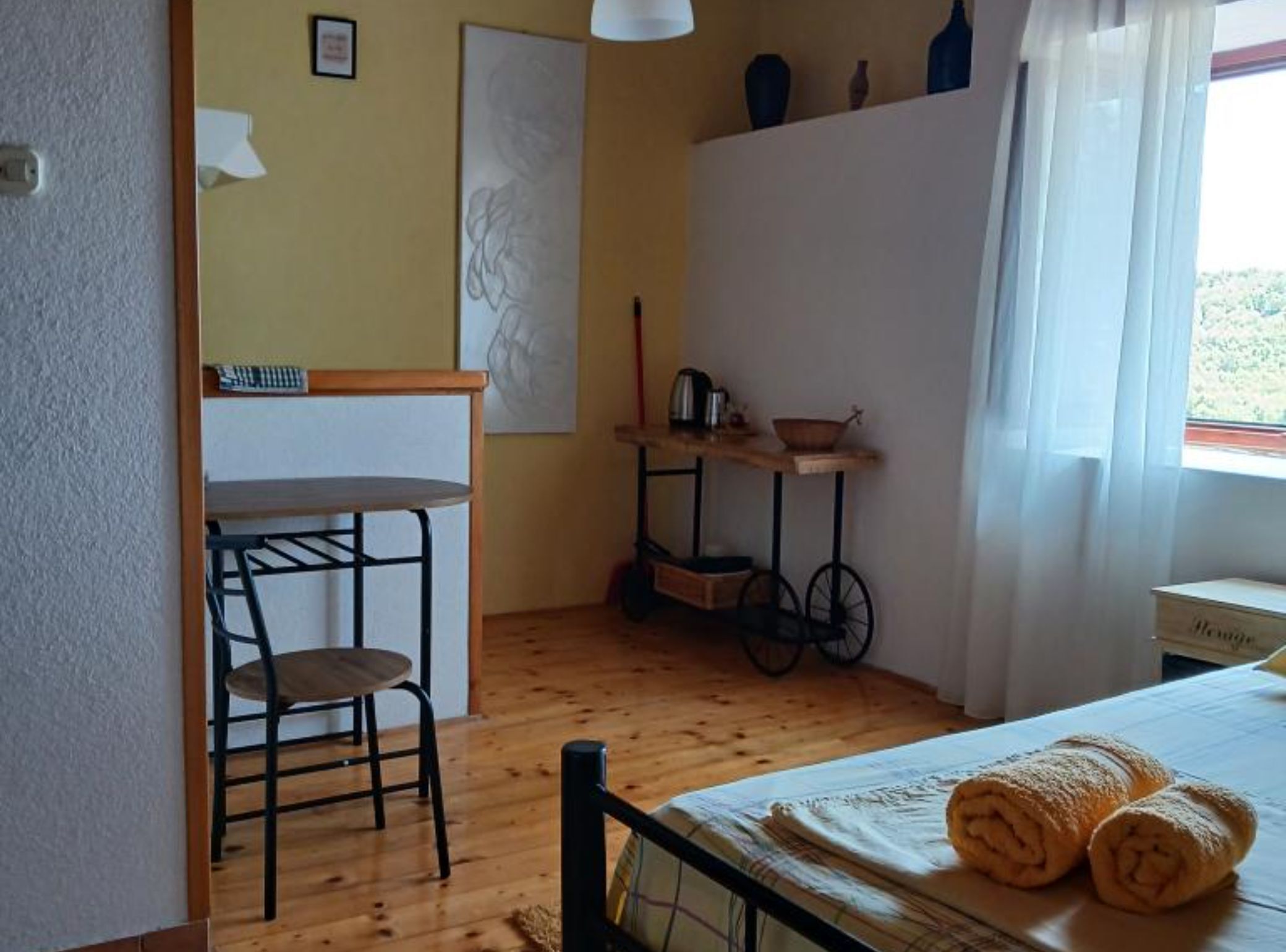 A small, cozy room with a wooden floor, a bed with rolled towels, a small table with a chair, a shelf with decorative items, and a window with curtains.