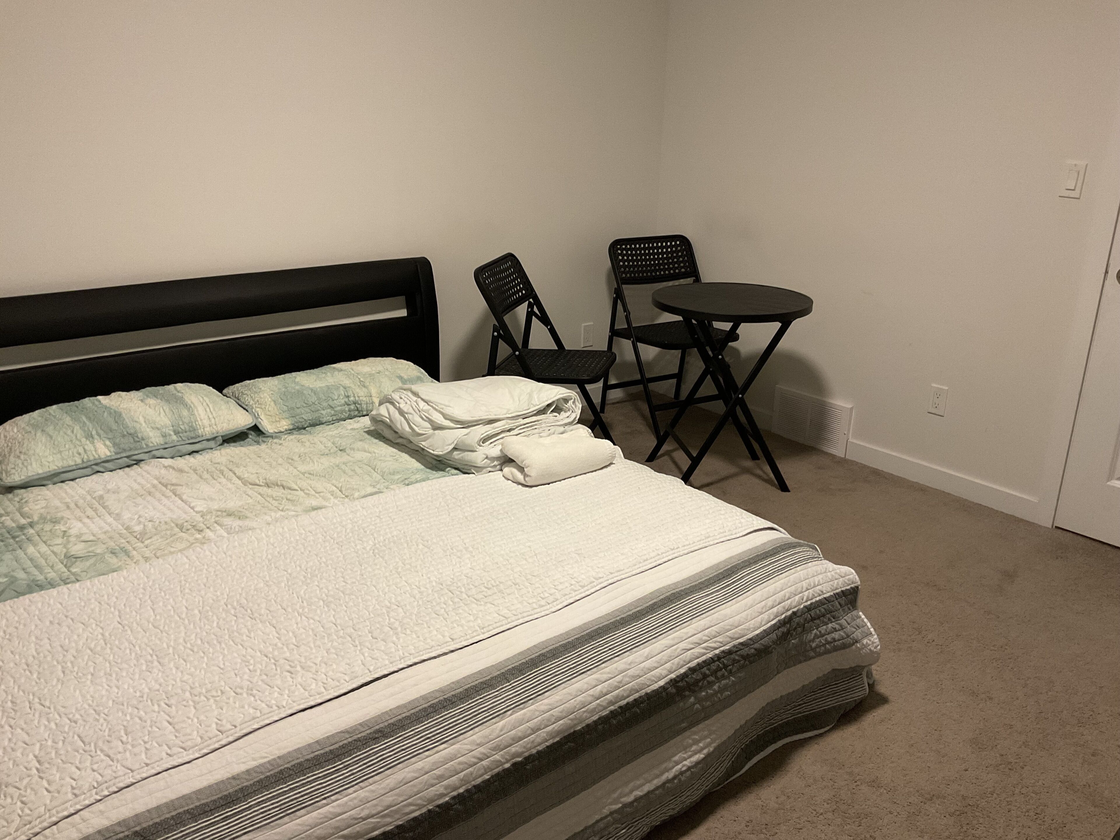 A neatly made bed with pillows and a folded blanket, a small black folding table with a chair, and a plain white wall with a power outlet.