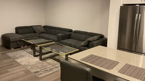 A modern living room with a sectional sofa, a glass-top dining table, and a stainless steel refrigerator.