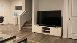 A modern living room with a flat-screen TV on a white stand, a staircase, and a water dispenser on the wall.