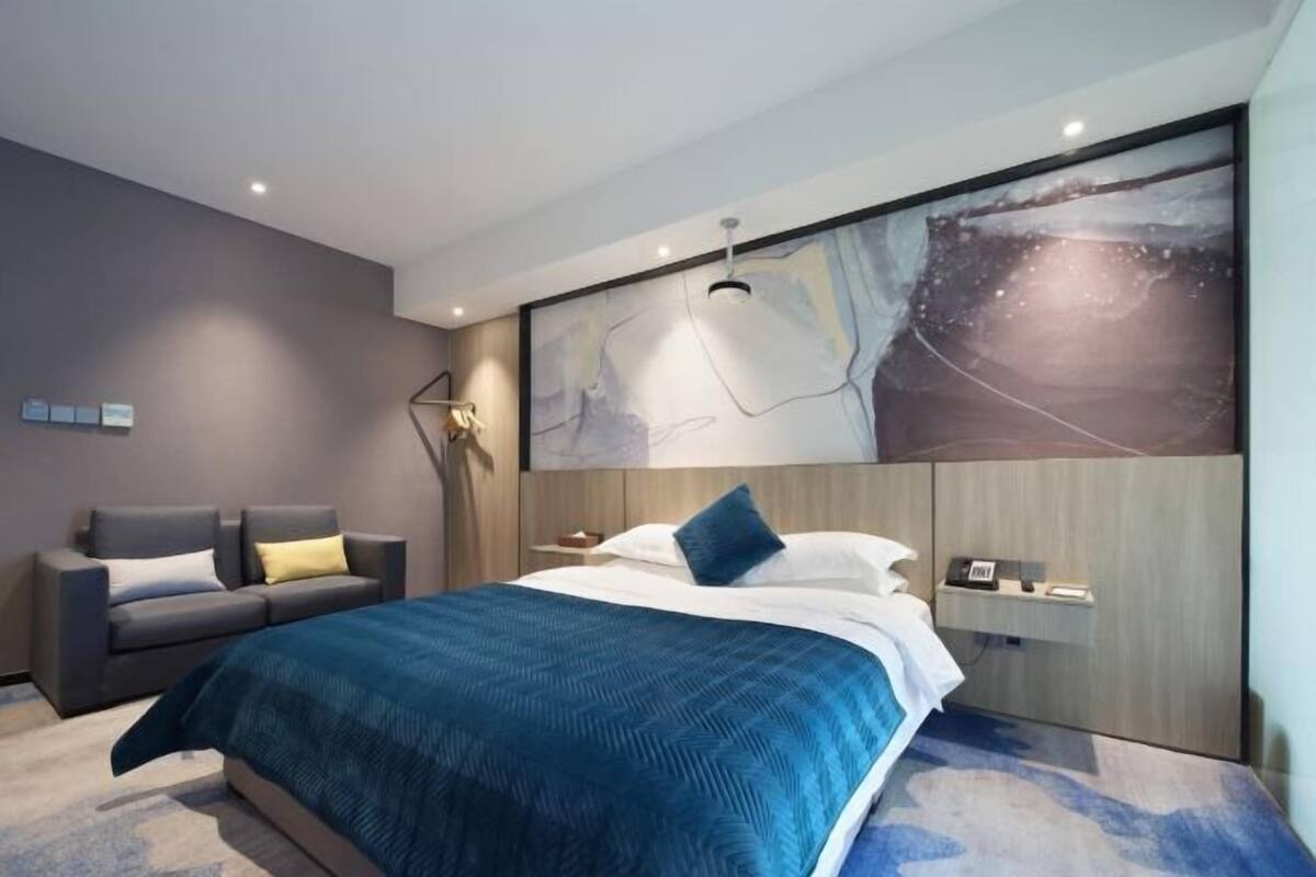 Une chambre d’hôtel moderne, équipée d’un lit, d’un canapé, d’un bureau et ornée d’un grand tableau abstrait au mur.