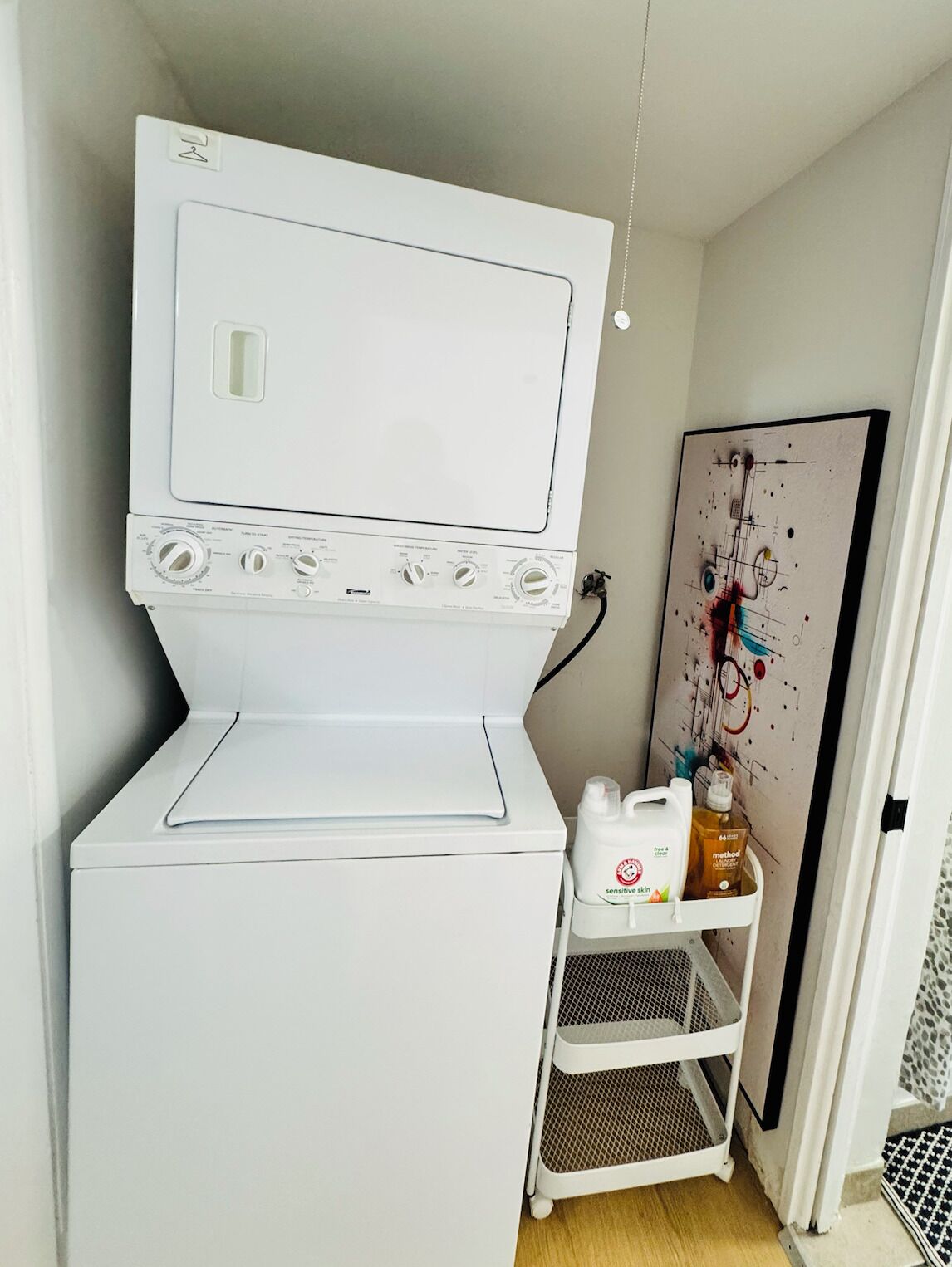 A compact laundry room with a front-loading washer and dryer, a small shelf with cleaning supplies, and a wall-mounted artwork.