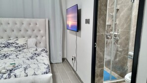 Ett sovrum med en stoppad sänggavel, en säng med ett blommigt överkast, en platt-TV som är monterad på väggen och en duschhörna med glasväggar.