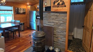 Interior rústico de una cabaña con chimenea de piedra, paredes de madera, comedor y cocina con refrigerador.