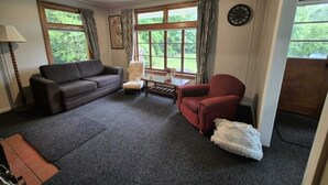 Una sala de estar con un sofá, un sillón y una mesita. Hay ventanas con cortinas, un reloj en la pared y una lámpara.