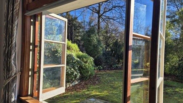 Vista a través de una ventana con marco de madera y múltiples vidrios, que da a un jardín con terraza de madera, plantas diversas y árboles.