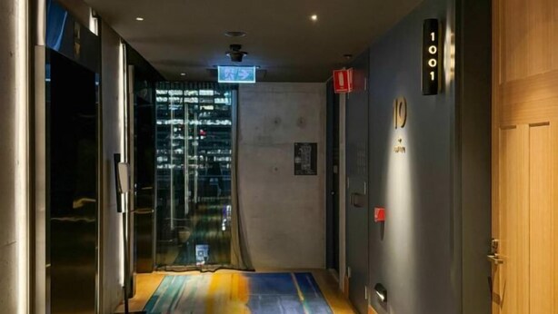 A hallway with a glass elevator, a door marked "1001", and a sign indicating the direction to the elevator. - Luxury Melbourne CBD Apt in Iconic QT Hotel (Melbourne)