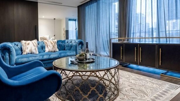 A modern living room with a blue sofa, a glass coffee table, and a view through large windows. - Luxury Melbourne CBD Apt in Iconic QT Hotel (Melbourne)
