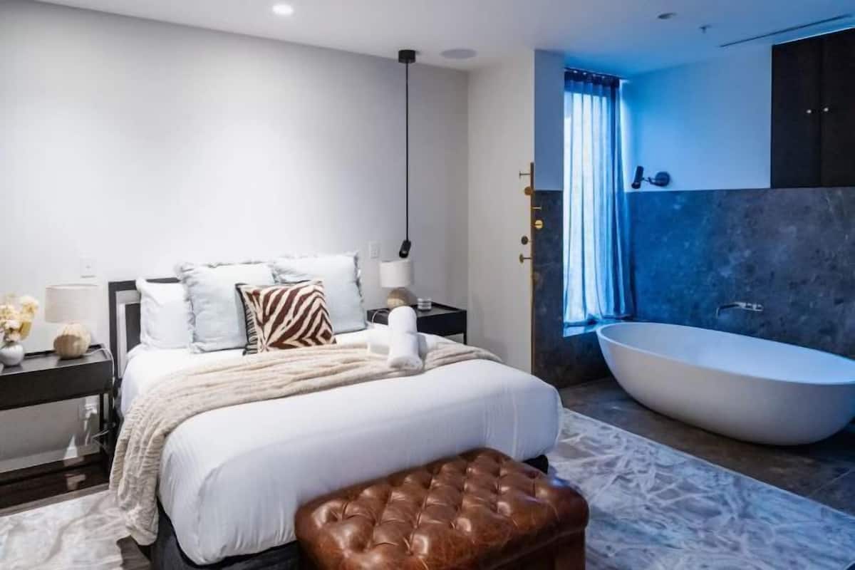 Ein modernes Schlafzimmer mit einem großen Bett, Nachttischen, einer Nachttischlampe, einem braunen Lederhocker und einer geräumigen Badewanne.