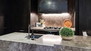 A modern kitchen with dark cabinetry, a stainless steel range hood, a marble countertop, a sink, and a potted plant. - Luxury Melbourne CBD Apt in Iconic QT Hotel (Melbourne)