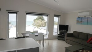 A modern living room with a flat-screen TV, a sofa, and a dining area with a table and chairs. Large windows offer a view of the sea and a building with a red roof. - Ultra-Modern Coastal Living (Apollo Bay)