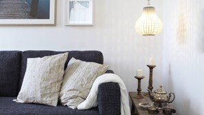 A cozy living room with a dark grey sofa, floral cushions, a white throw blanket, a wooden side table with candles and a teapot, and a hanging light fixture. - KÖNIGSschlaf's large vacation apartment for up to 3-5 people with sunny balcony (Konstanz)