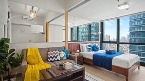 A modern hotel room with a large bed, a sofa, a coffee table, and a large window offering a city view. - BGC 1BR, Steps to Mitsukoshi & Uptown Malls, 300Mbps Fiber 19C2 (Taguig)