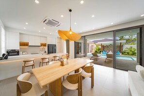 A modern kitchen with white cabinets, a wooden dining table, and a large window overlooking a pool area. - Valentine Village (Koh Samui)